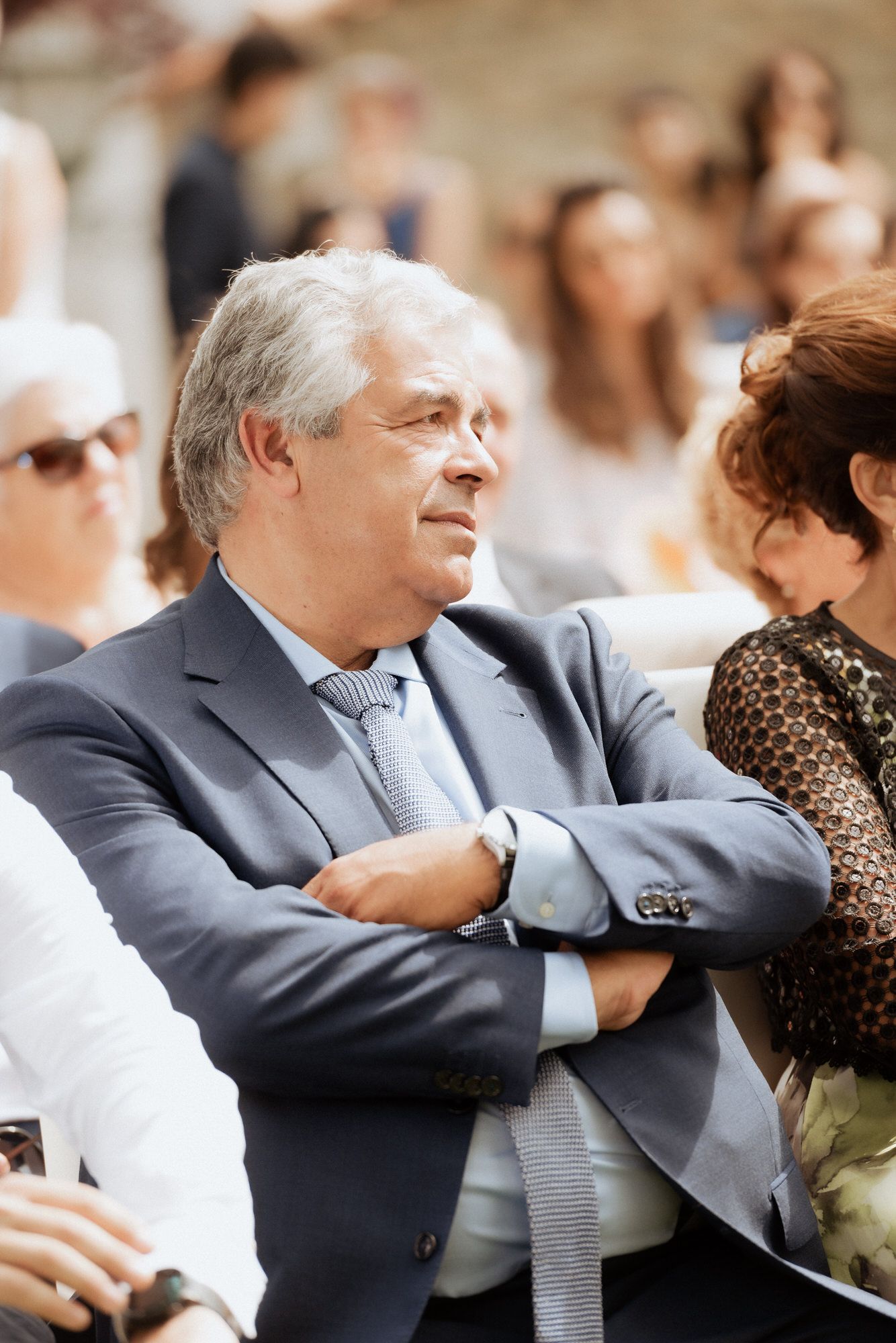 A grey-haired person in a suit stands with arms crossed among other attendees at an outdoor event.
