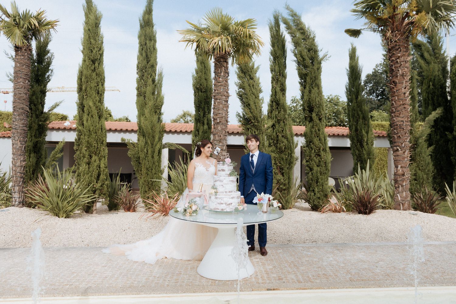 A couple stands next to their elegant white wedding cake on a pedestal with tall cypress trees and palm trees in the background.