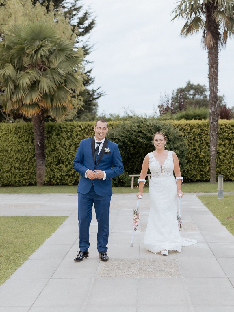 A wedding couple walks along a path bordered by palm trees and manicured hedges on a sunny day.