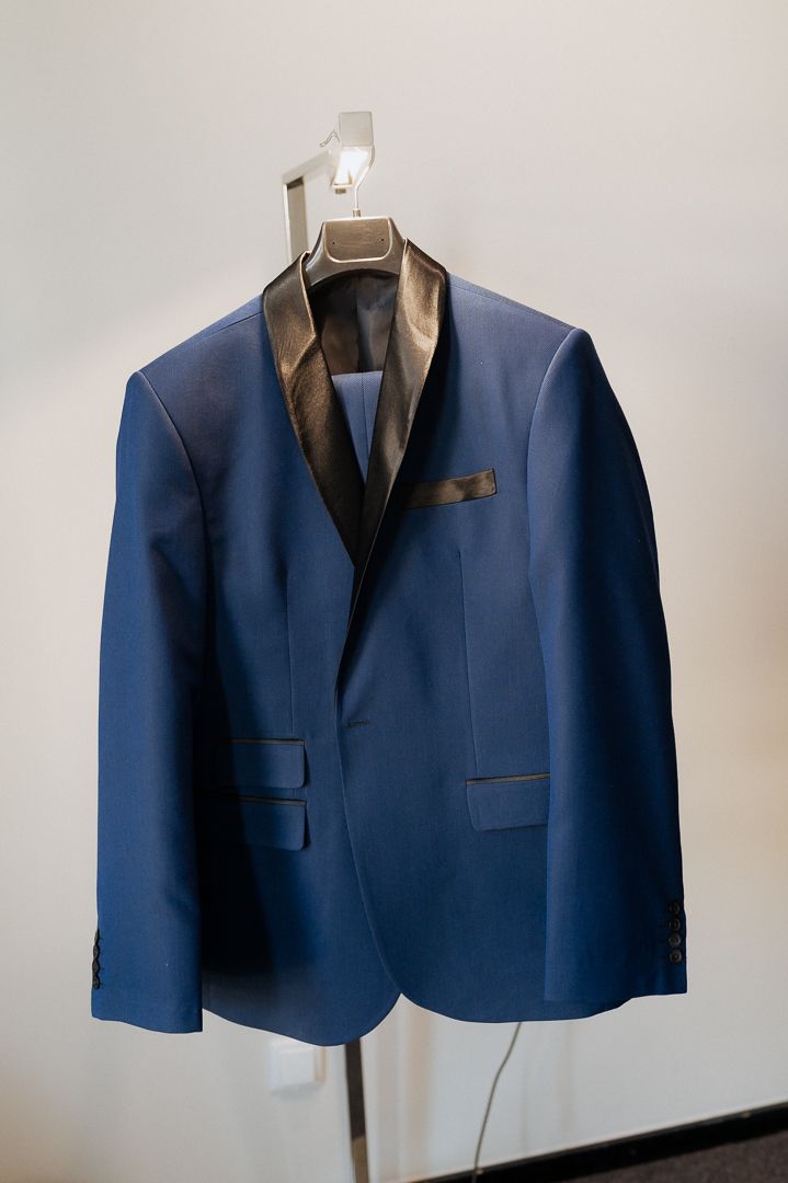 Navy blue tuxedo jacket with brown satin lapels hanging on white display hanger against light wall.