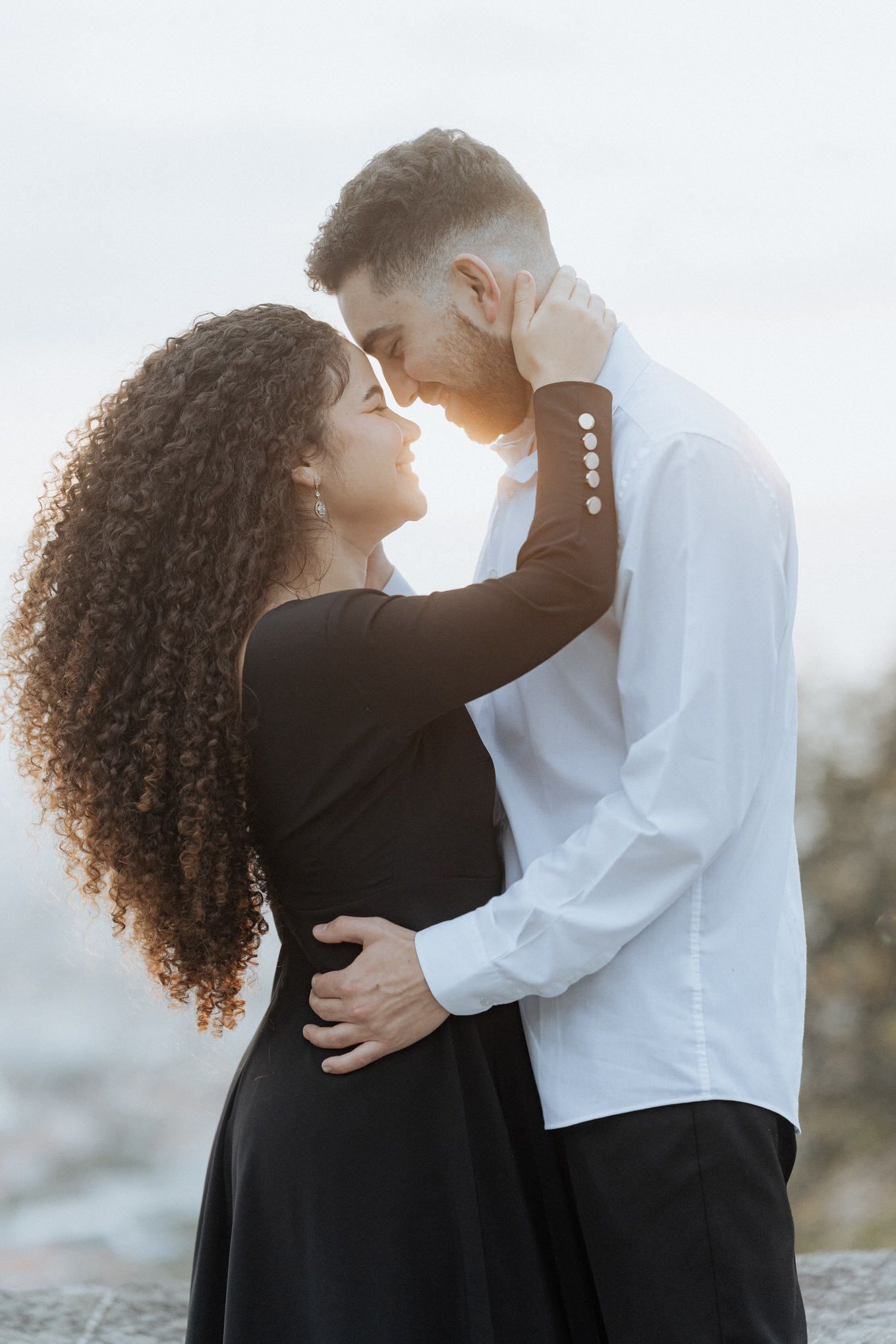 Casal romântico se abraça ao pôr do sol com um fundo enevoado, com o cabelo cacheado dela e o vestido preto contrastando com a camisa branca