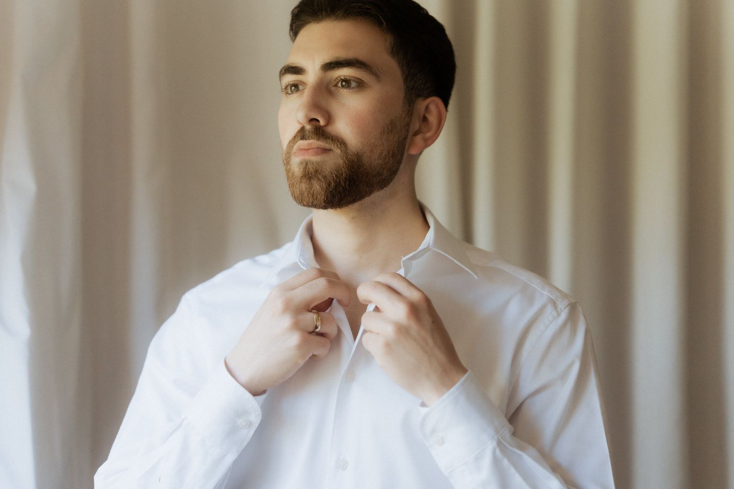 A person in a white button-up shirt adjusts their collar while standing against a light curtained background.
