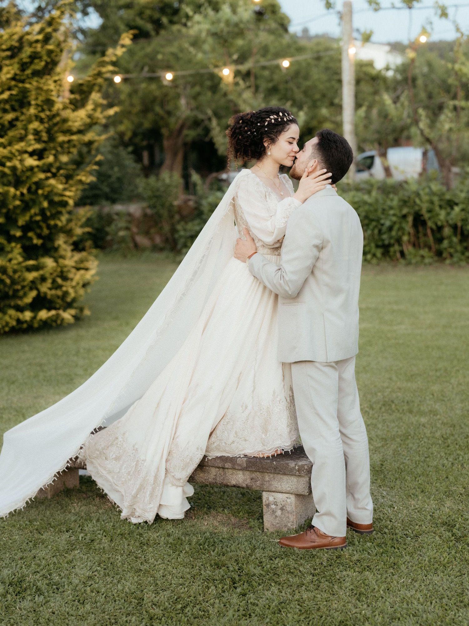A romantic outdoor wedding photograph showing a couple in cream-colored attire kissing on a rustic wooden block in a garden.