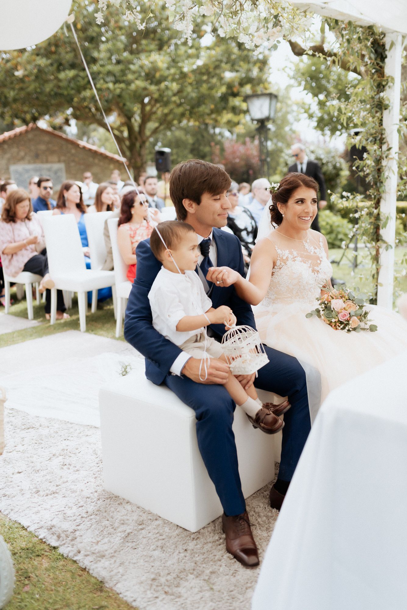 Bride in white dress and groom in navy suit sit together on outdoor white bench during garden reception ceremony.