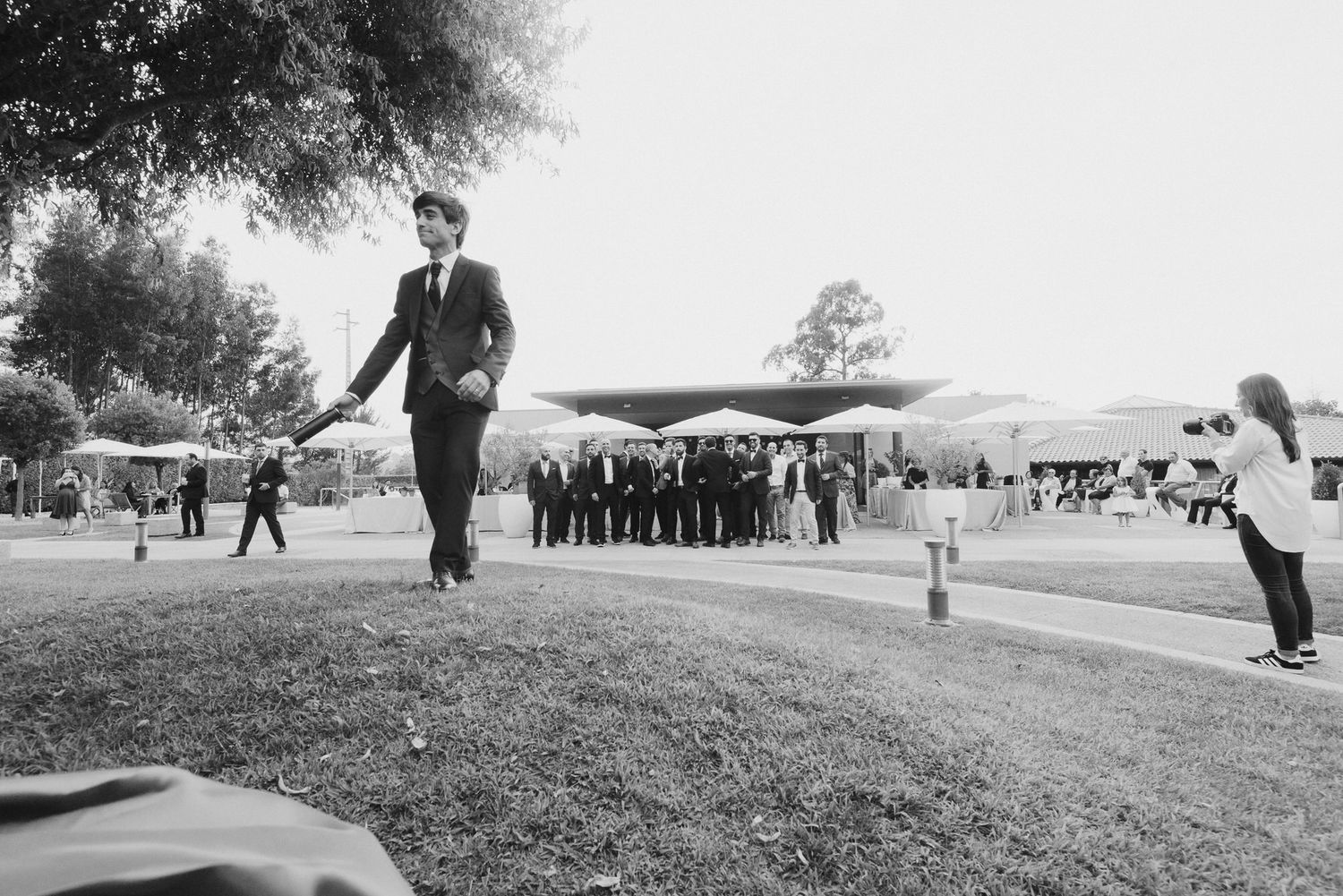 A black and white photograph showing someone walking along a grassy slope with a crowd gathered below in the distance.