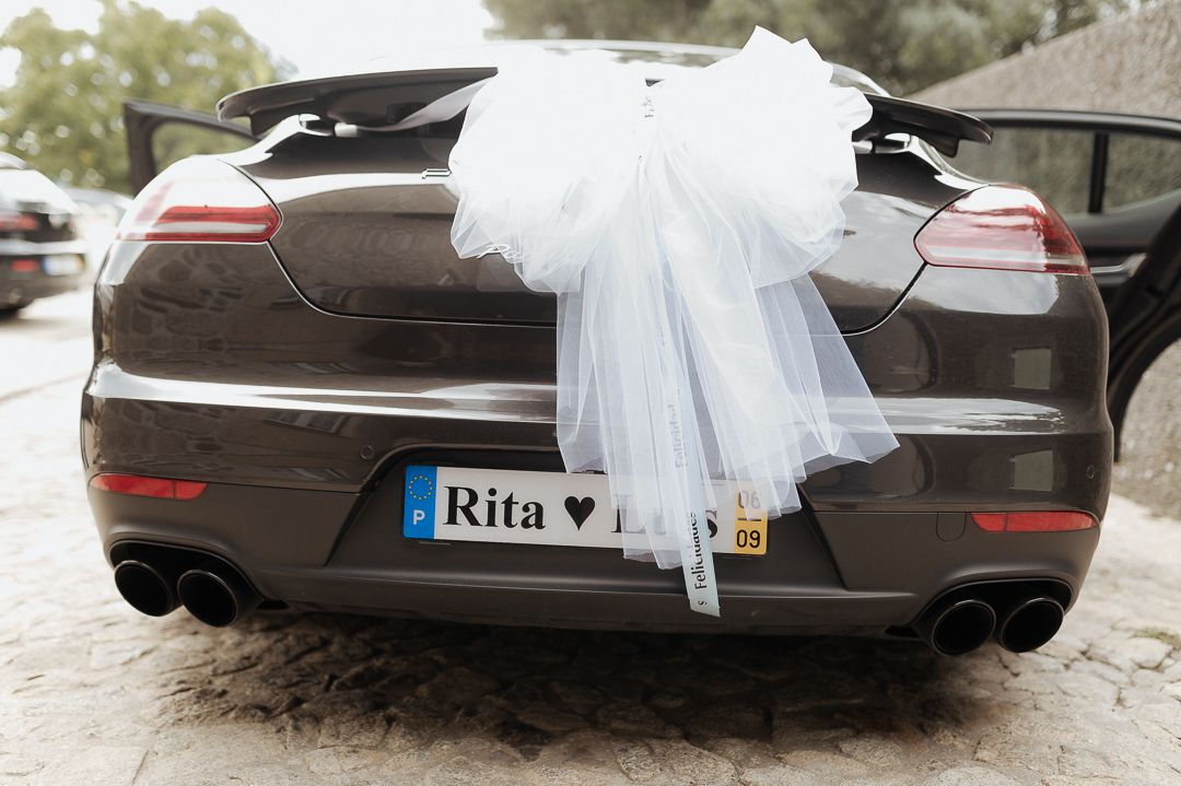 Black wedding car decorated with white tulle bow and personalized 'Rita' license plate shown from rear view.
