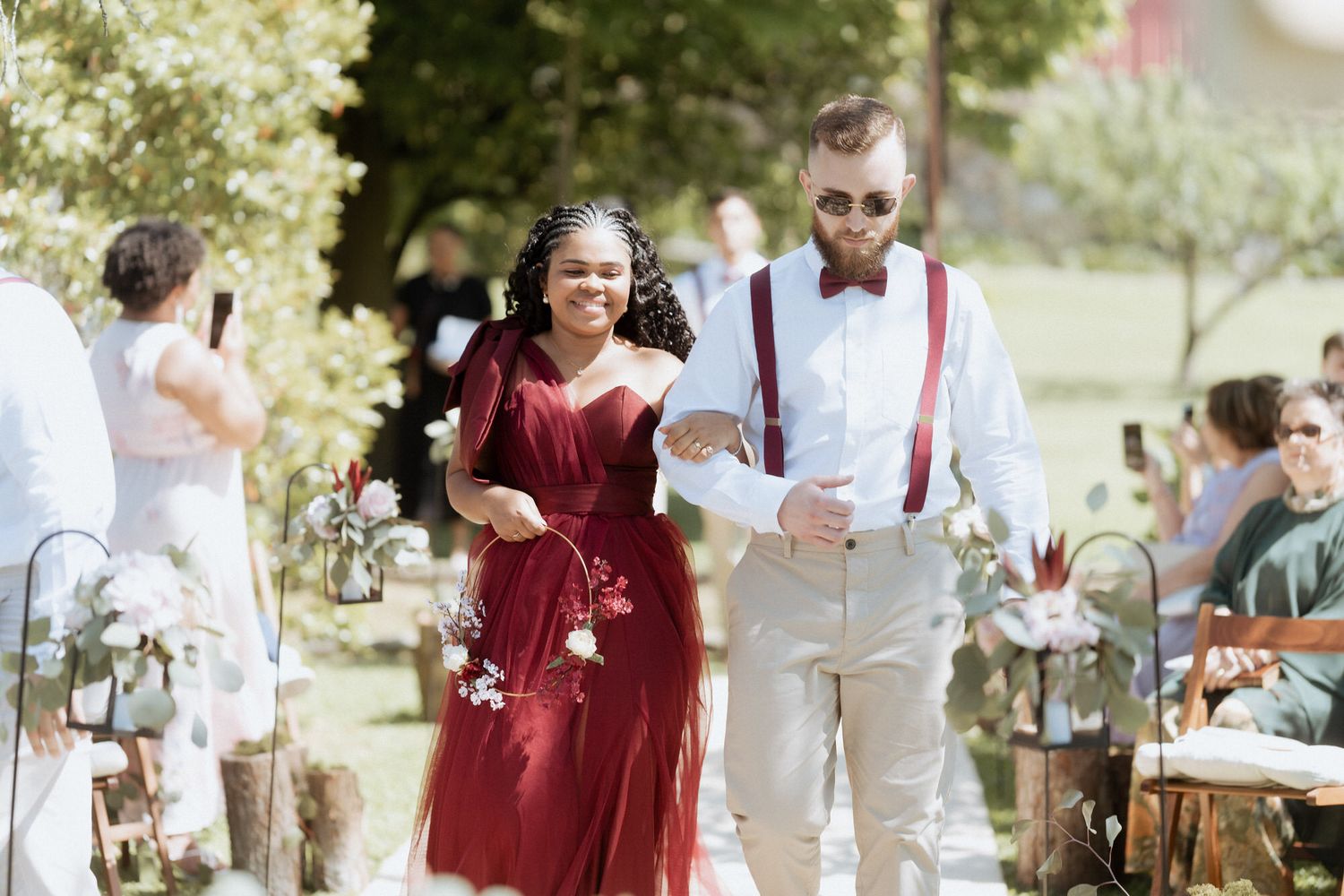 Wedding party member in burgundy dress walks down aisle with groomsman in white suit and suspenders at outdoor ceremony.