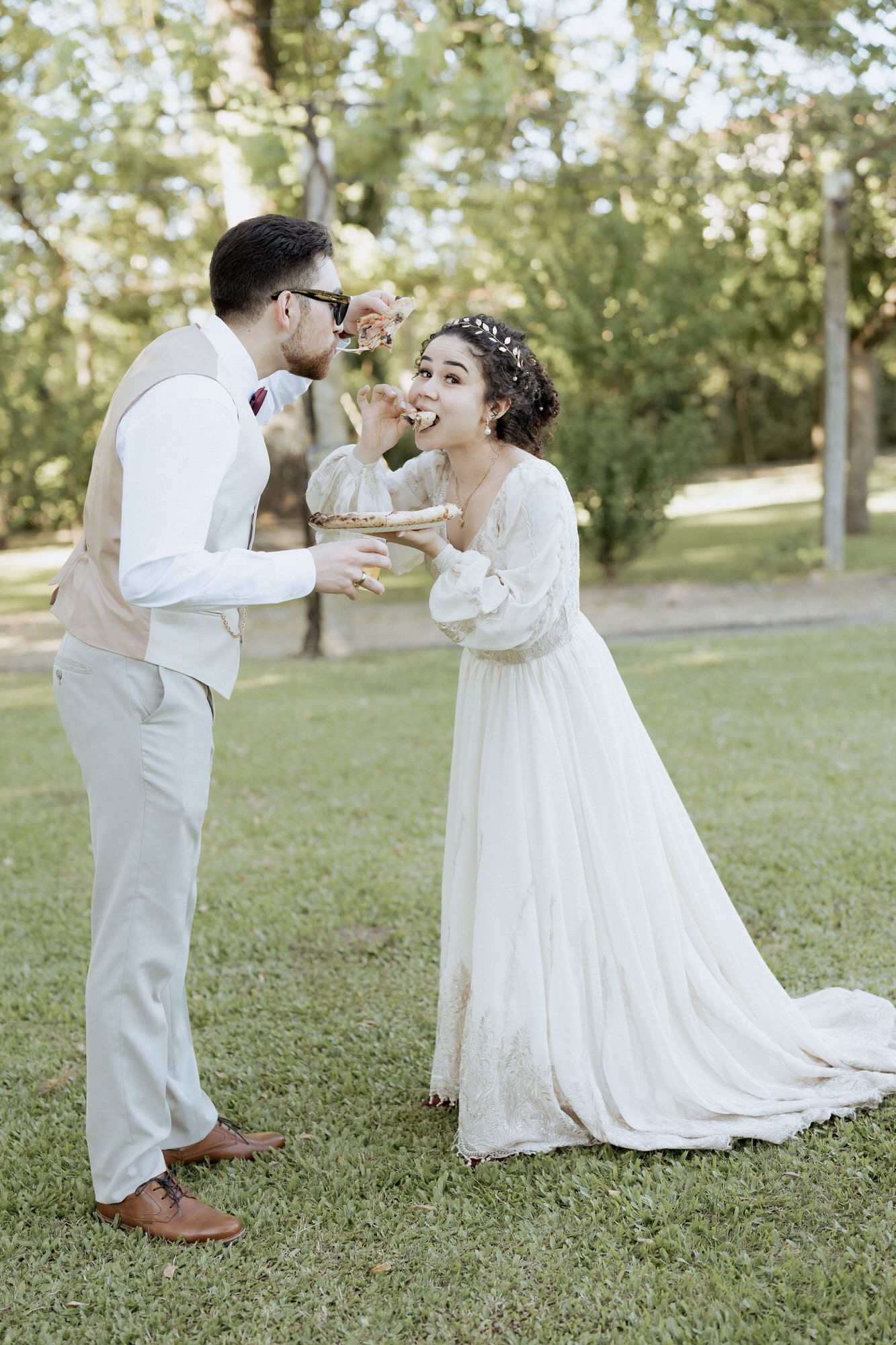 Newlywed couple in outdoor wedding attire playfully feed each other cake in a sunny garden setting.