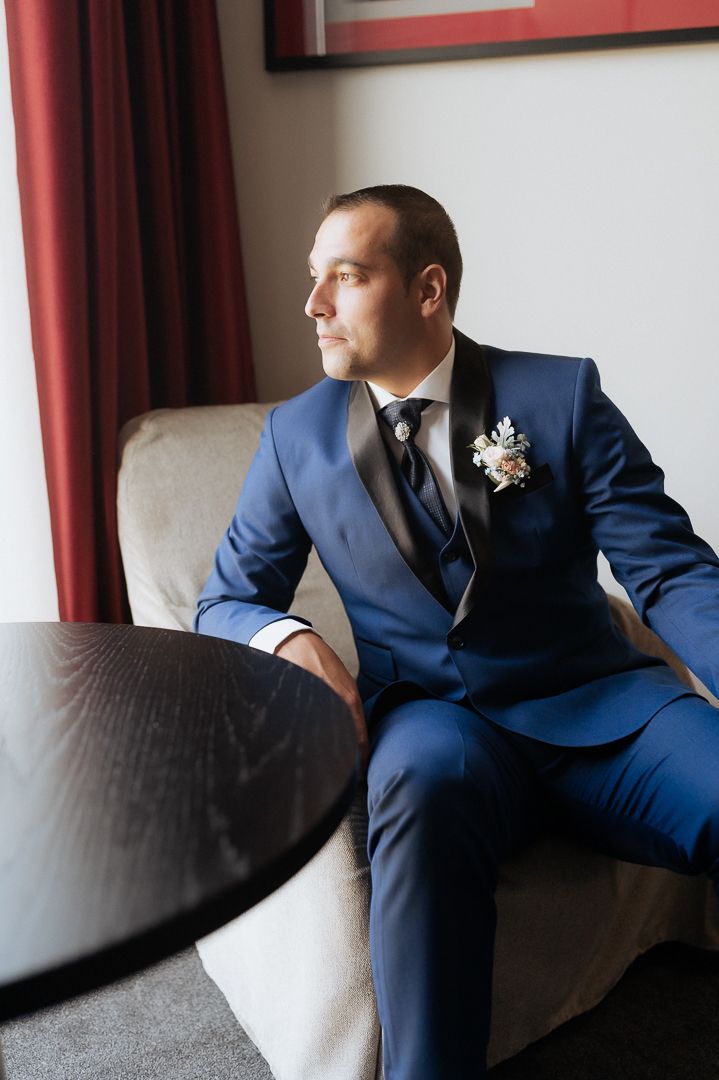 A groom in a navy blue suit sits thoughtfully by a window with a boutonniere pinned to his lapel.