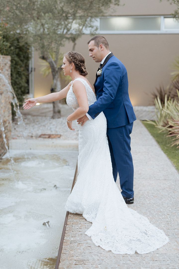Couple in formal wedding attire walking together on path with white lace dress train trailing behind.