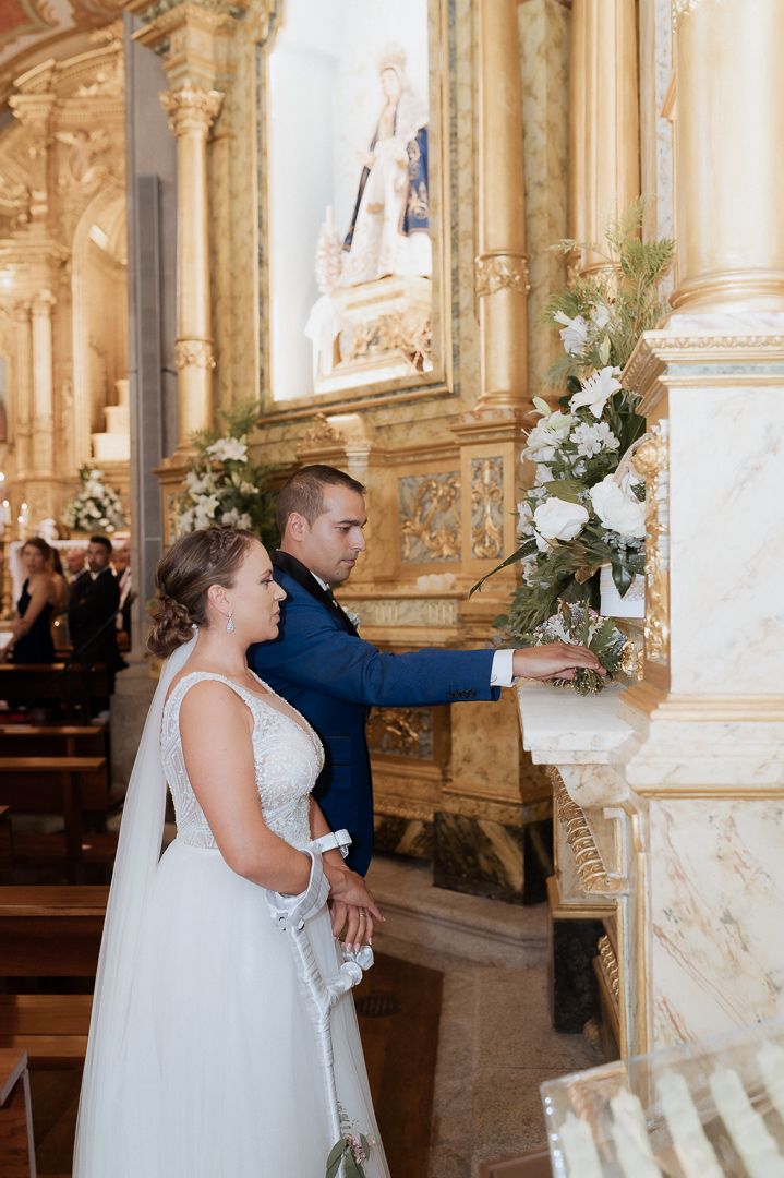A couple in wedding attire stands before an ornate marble altar adorned with white floral arrangements in a grand church.