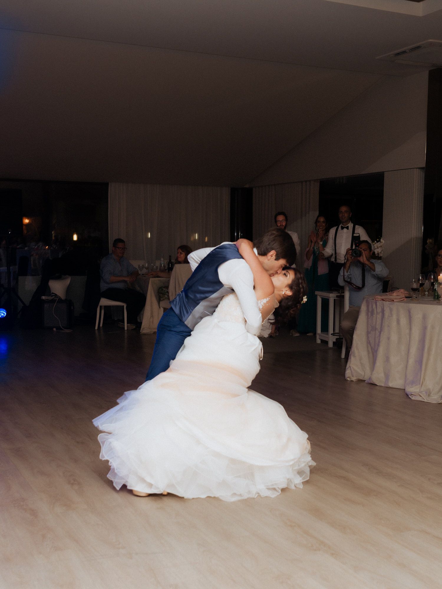 Newlyweds share a romantic dip dance on the wooden floor at their wedding reception with decorative lighting.