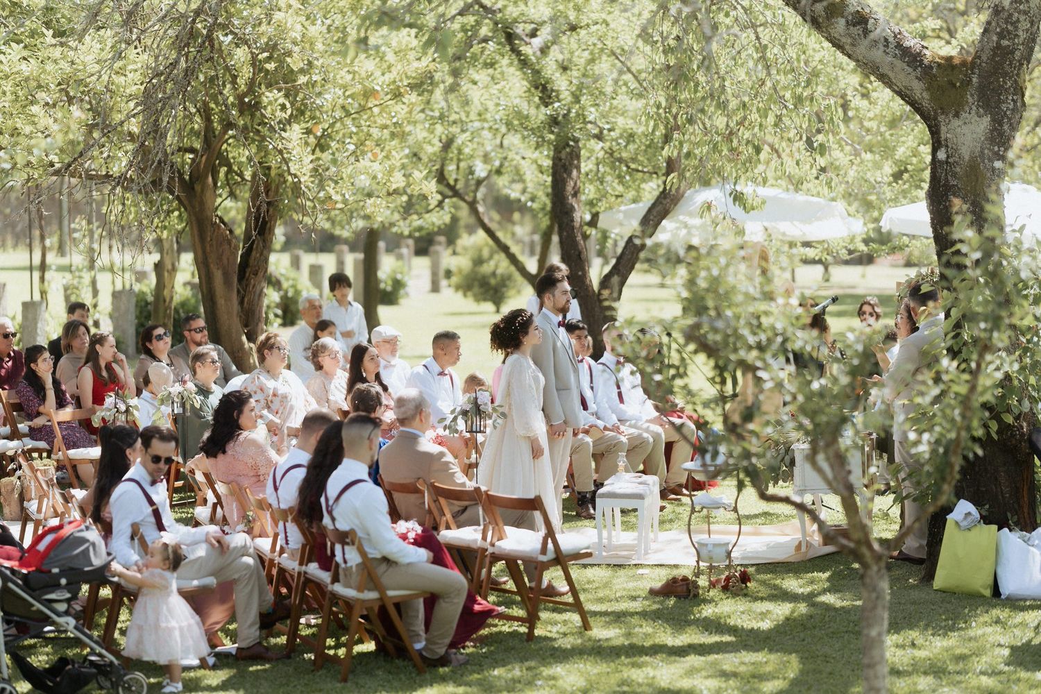 Outdoor garden wedding ceremony with guests seated on folding chairs under shady trees on a sunny summer day.