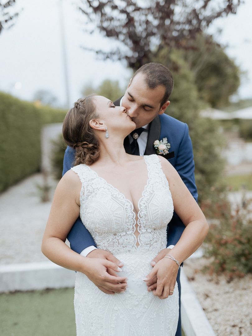 A romantic wedding couple embrace outdoors, with the bride in a lace gown and groom in a navy suit with boutonniere.