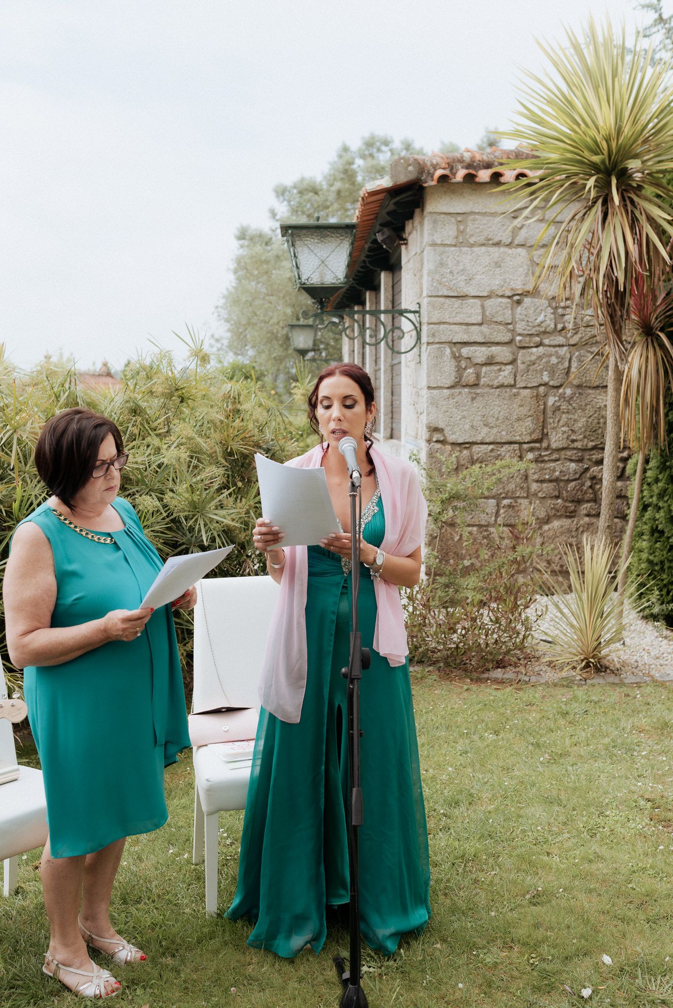 Two women in green dresses stand outside reading papers during an outdoor event at a stone wall garden.