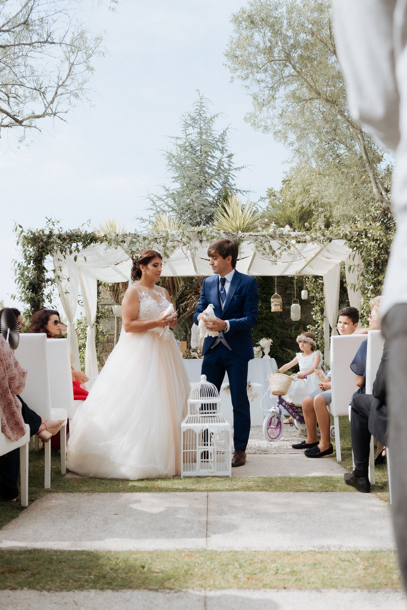 A romantic outdoor wedding ceremony under a white draped arch with a decorated aisle and natural garden setting.