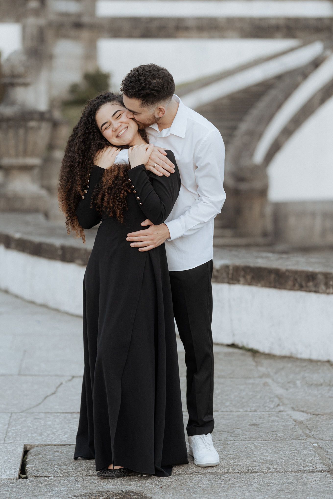 A couple embraces tenderly on a stone pathway while wearing formal black and white attire.