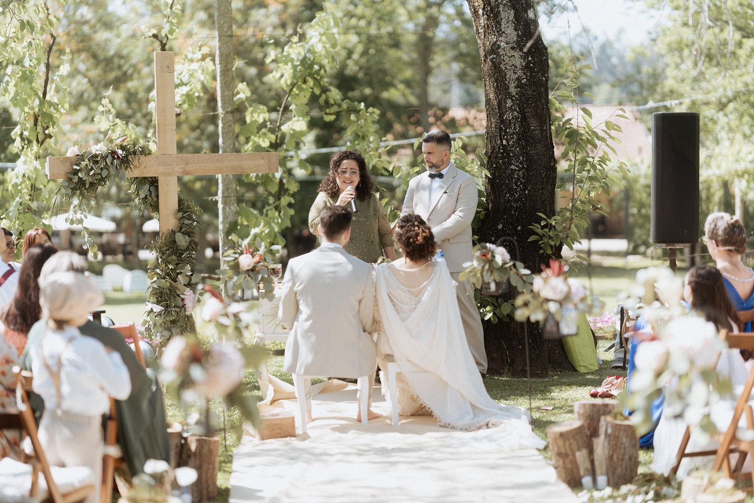 Outdoor wedding ceremony in a sunlit garden setting with white chairs and floral decorations along a white aisle runner.