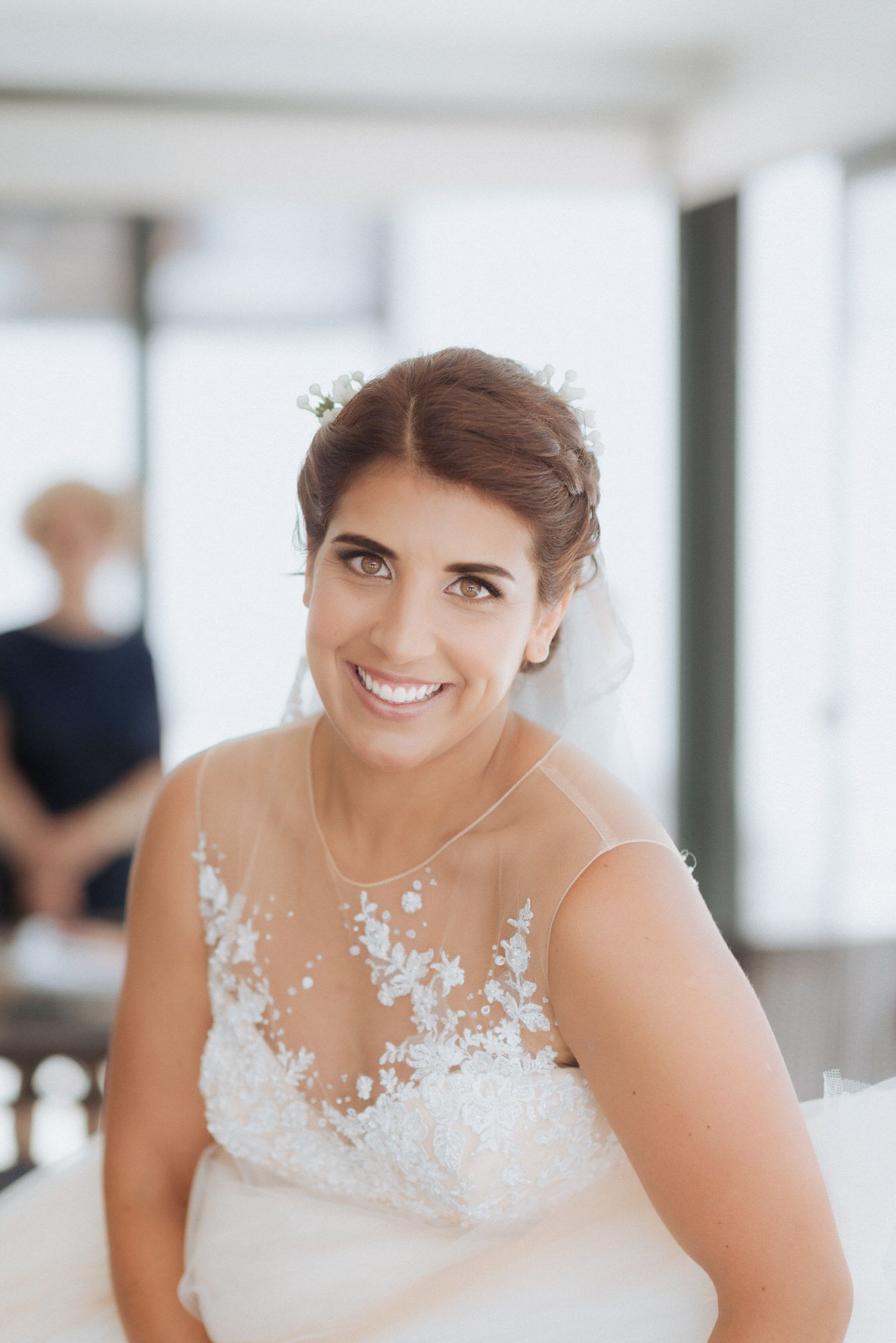 A bride in an illusion neckline gown with lace detail smiles warmly while preparing for her wedding day.