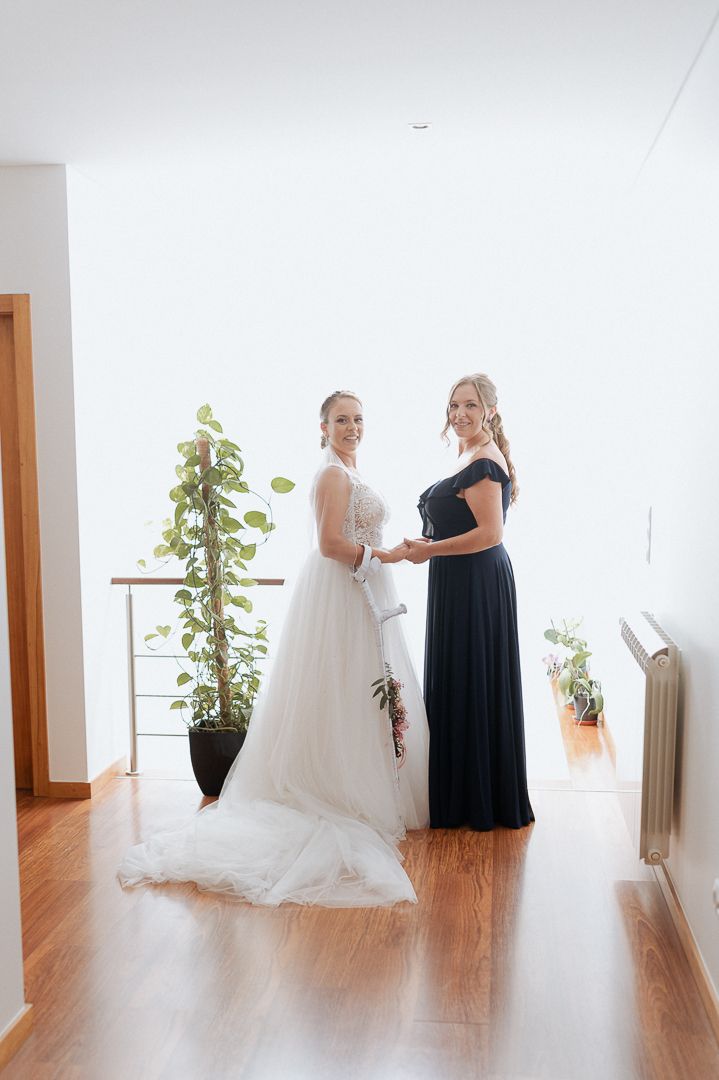 Two women stand in an elegantly lit room, one wearing a white wedding gown and the other in a navy evening dress.