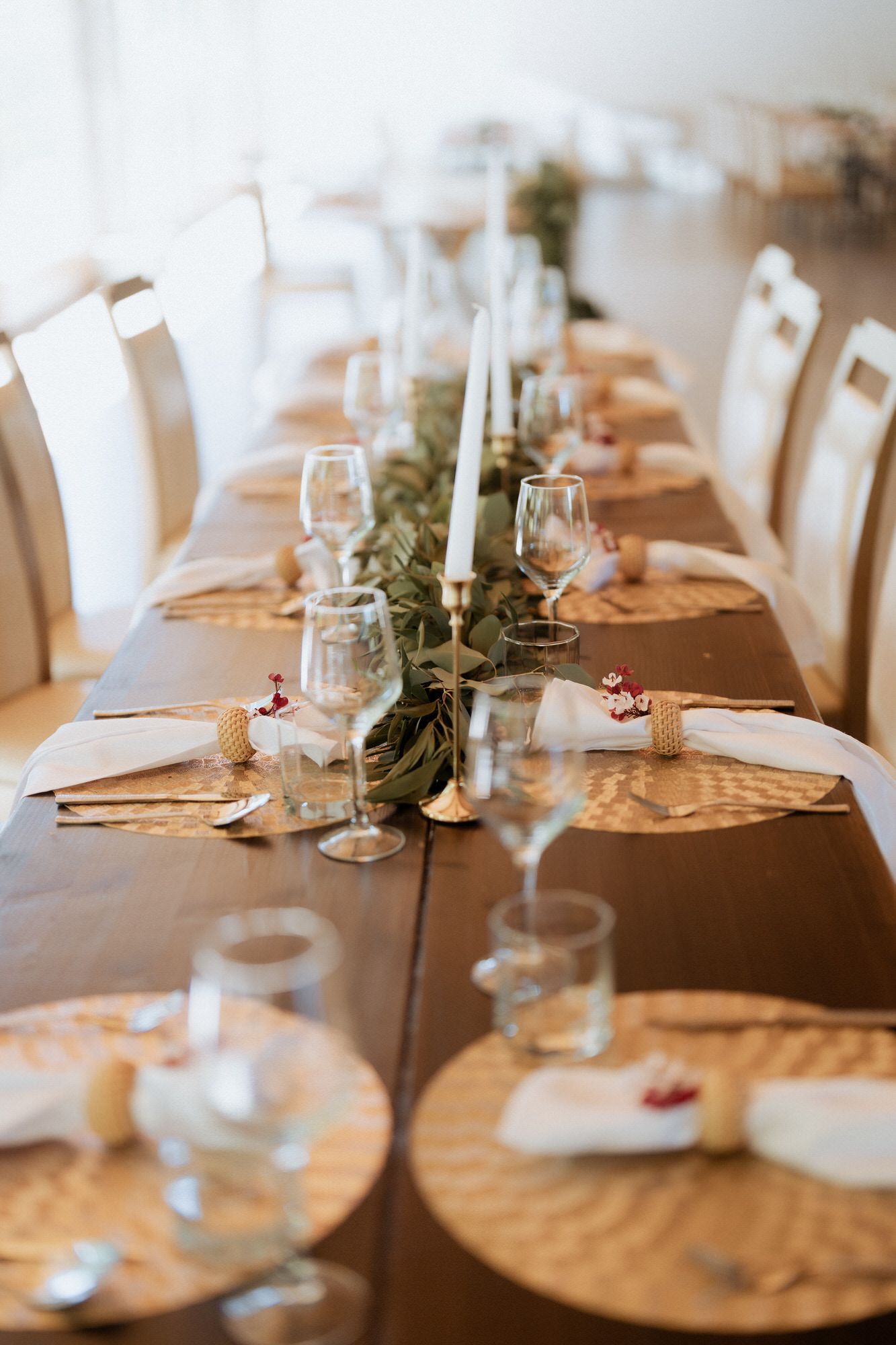 Elegant table setting with white candles, greenery runner and place settings on a brown tablecloth for a formal dinner.