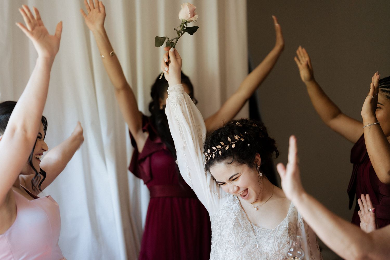 A group of bridesmaids in burgundy dresses and a bride in white raise their hands in celebration.