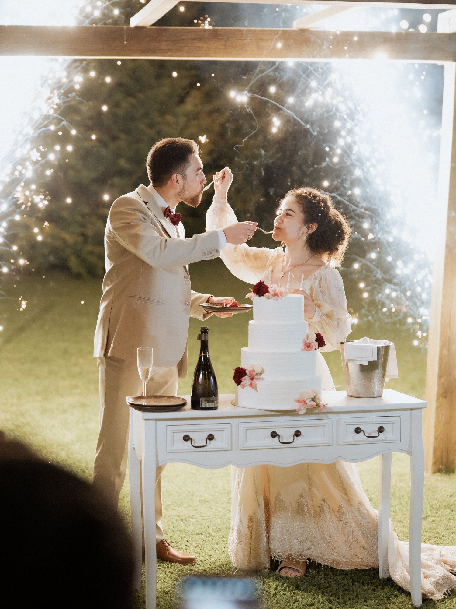 Joyful couple shares a cake-tasting moment at their outdoor wedding reception by a vintage white table.