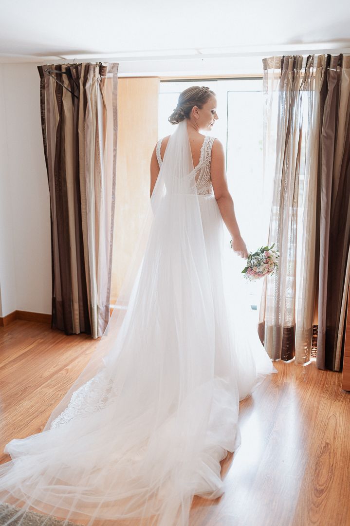 Elegant white wedding gown with flowing train and lace detail shown from behind near brown curtained windows.