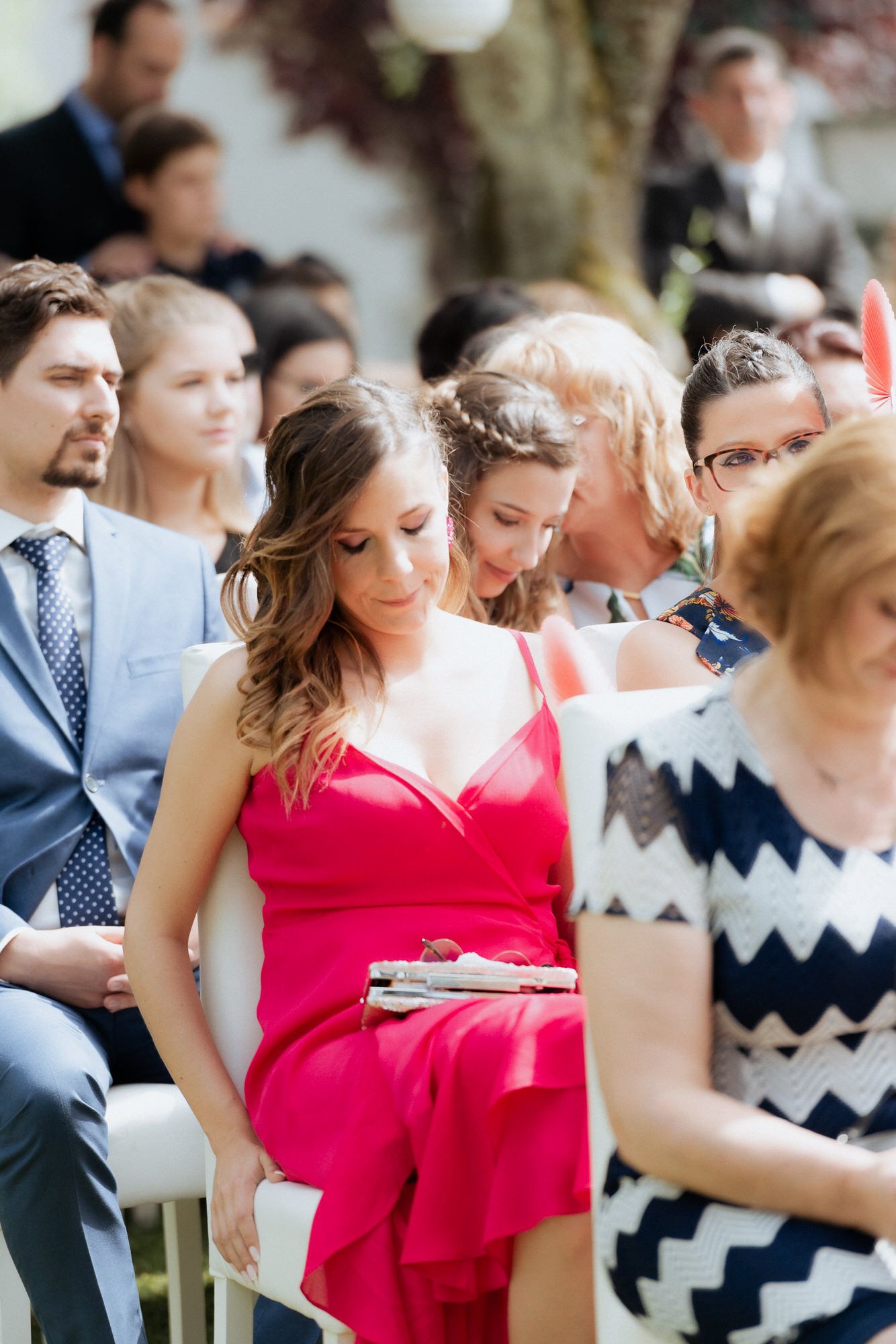 Wedding guests in formal attire seated at an outdoor ceremony, with a woman in a vibrant red dress in focus.