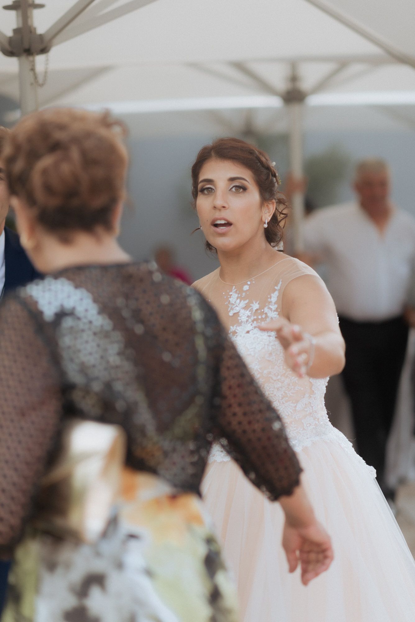 A bride in a white lace wedding gown gestures while in conversation during an outdoor reception.