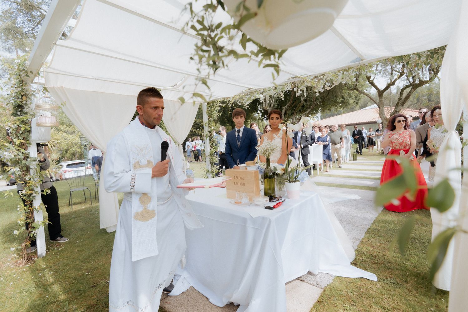 A wedding celebration takes place under an elegant white outdoor tent decorated with greenery and floral arrangements.