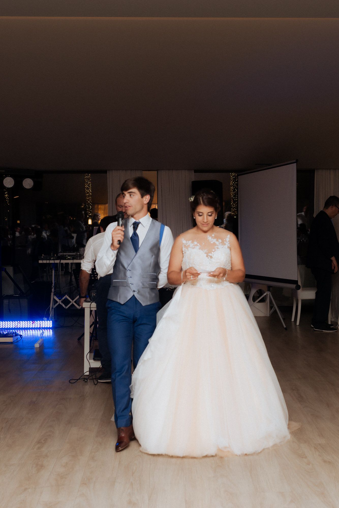 Two people stand together at a wedding reception, with the person in a blue suit speaking into a microphone and a bride in white.