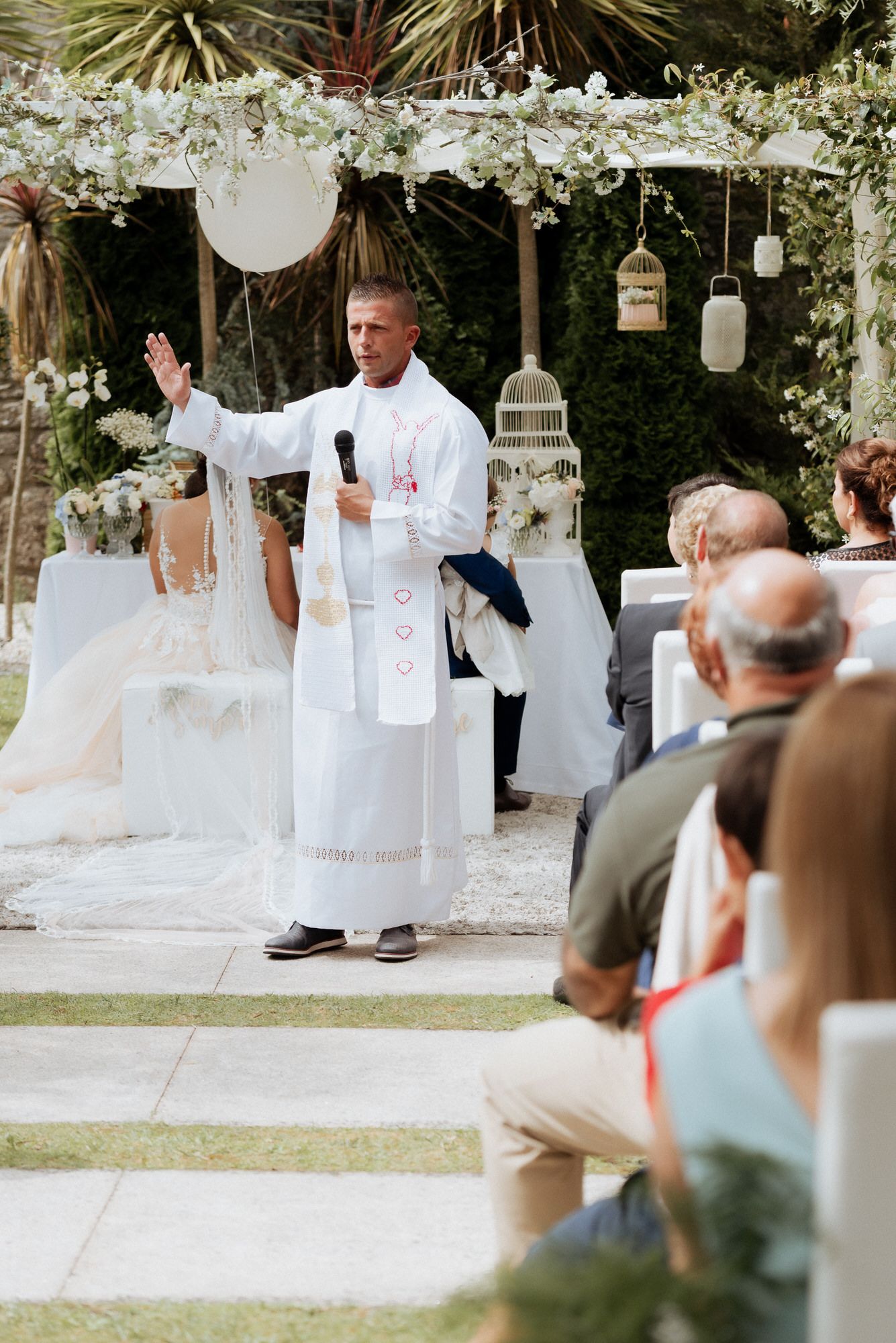 An outdoor wedding ceremony under a floral arch with hanging lanterns and decorative details.