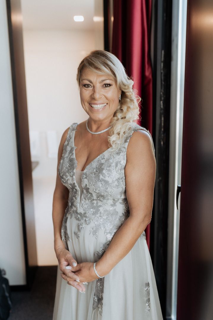 A bride wearing an elegant lace and tulle wedding gown smiles while posing in a room with red curtains.