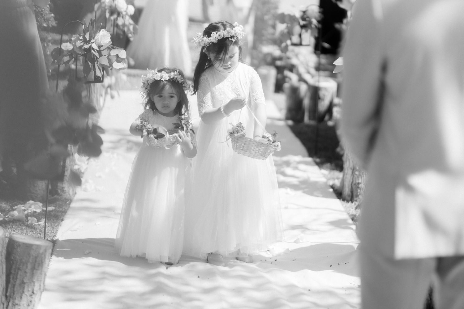A dreamy black and white photo shows flower girls in flowing white dresses walking along an outdoor wedding aisle.