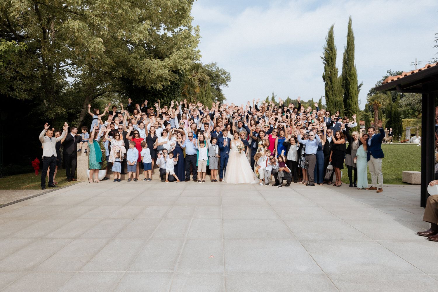 A large group of wedding guests gathered outdoors with cypress trees and landscaping in the background.
