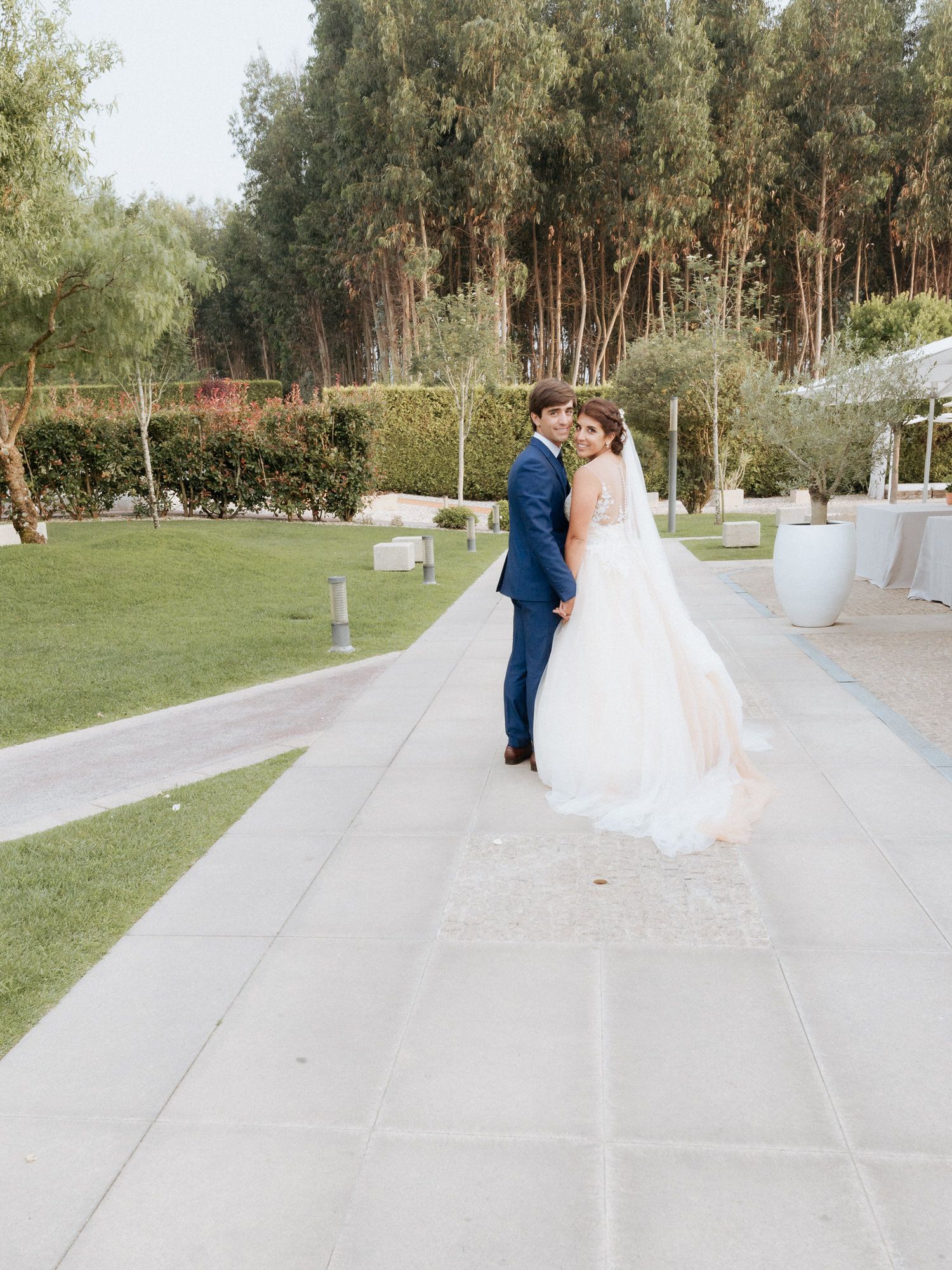 Elegant outdoor wedding sequence showing a couple in navy and white attire walking together on a stone pathway surrounded by trees.