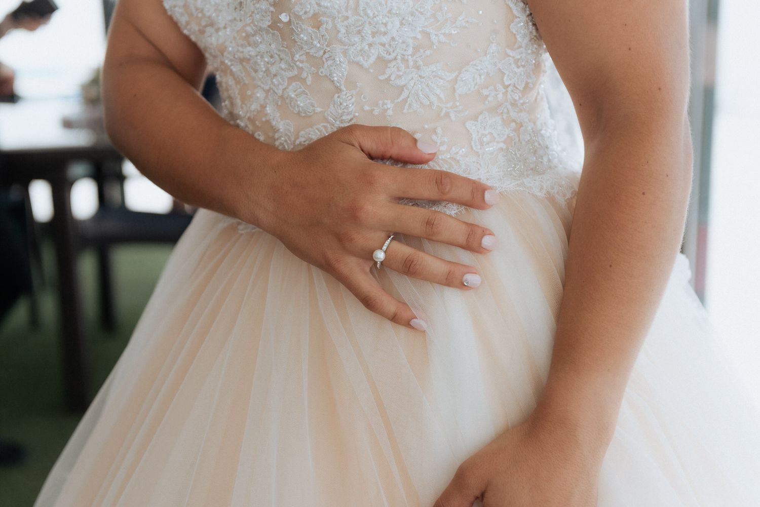 Close-up view of a bride's hand with engagement ring resting on her white lace and tulle wedding gown.