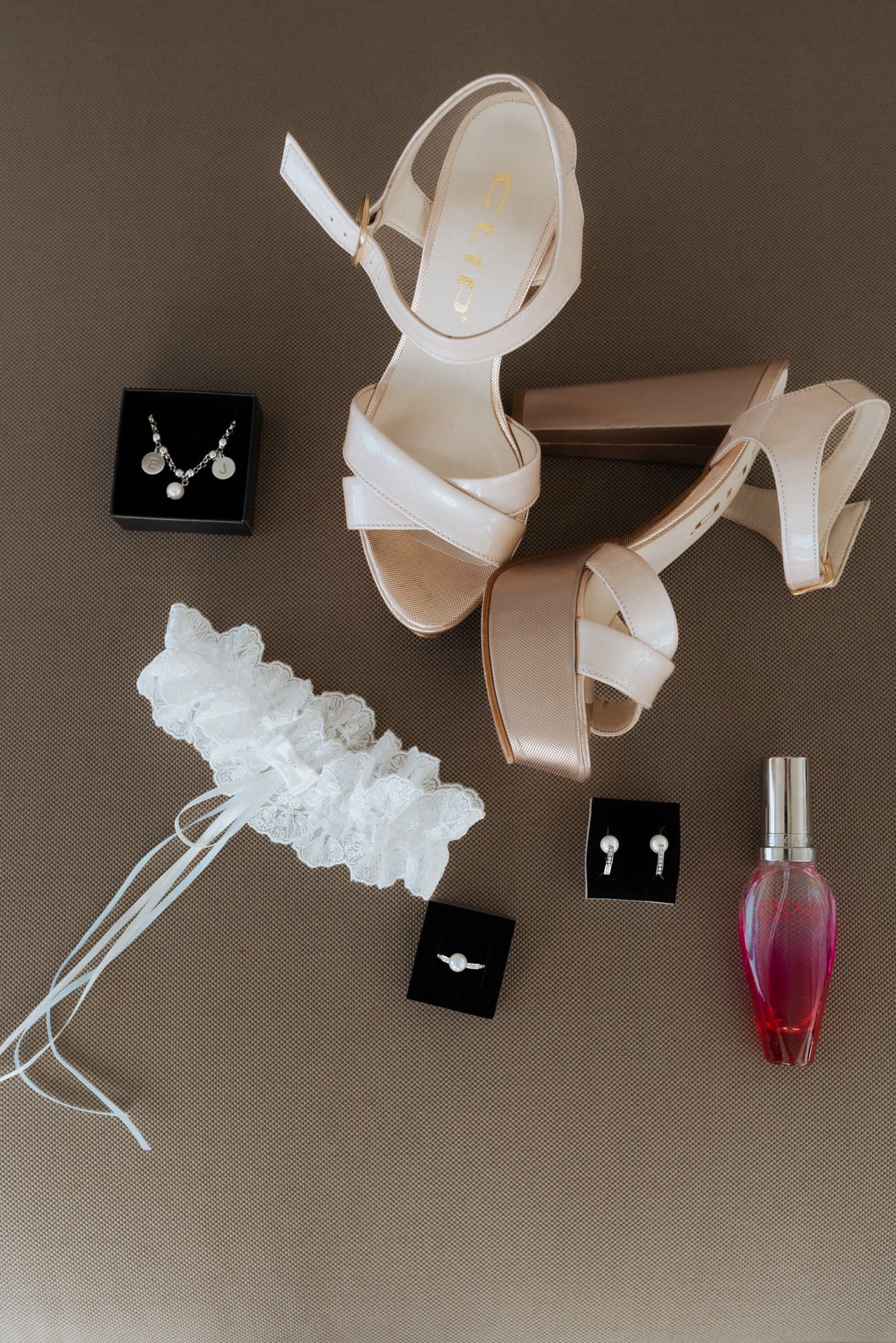Bridal accessories including white heels, garter, jewelry boxes, and pink perfume bottle arranged on beige surface.