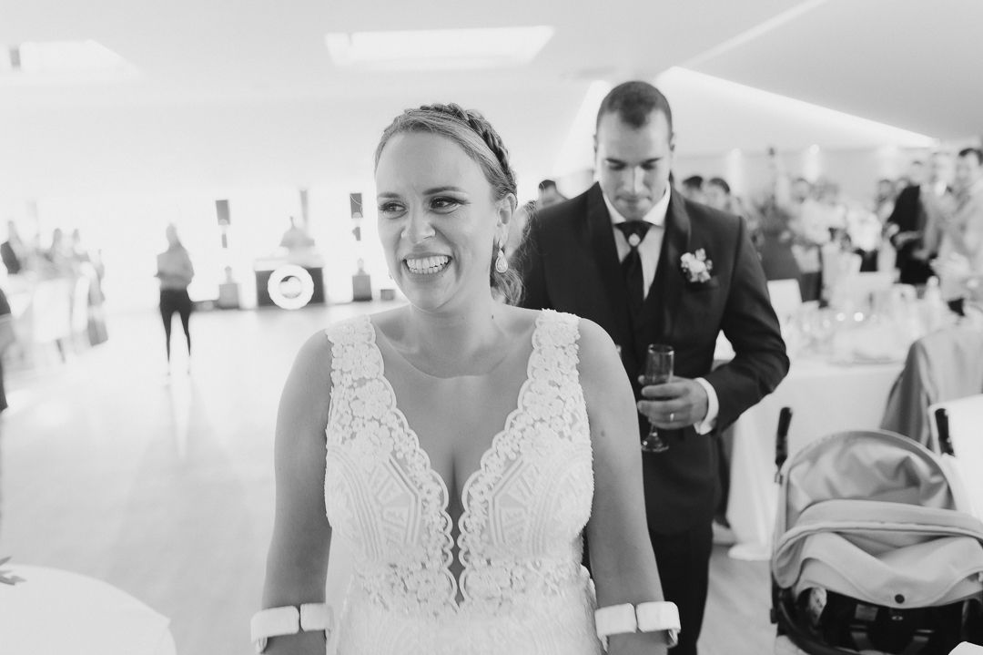 Black and white photo of bride in lace wedding dress with guest in formal wear in background at reception venue.