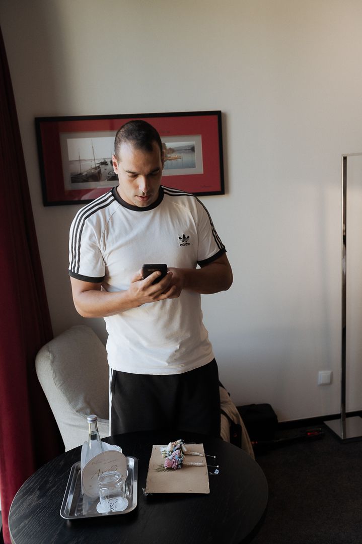 Person in white Adidas t-shirt checking phone while standing next to round table with drinks and snacks.