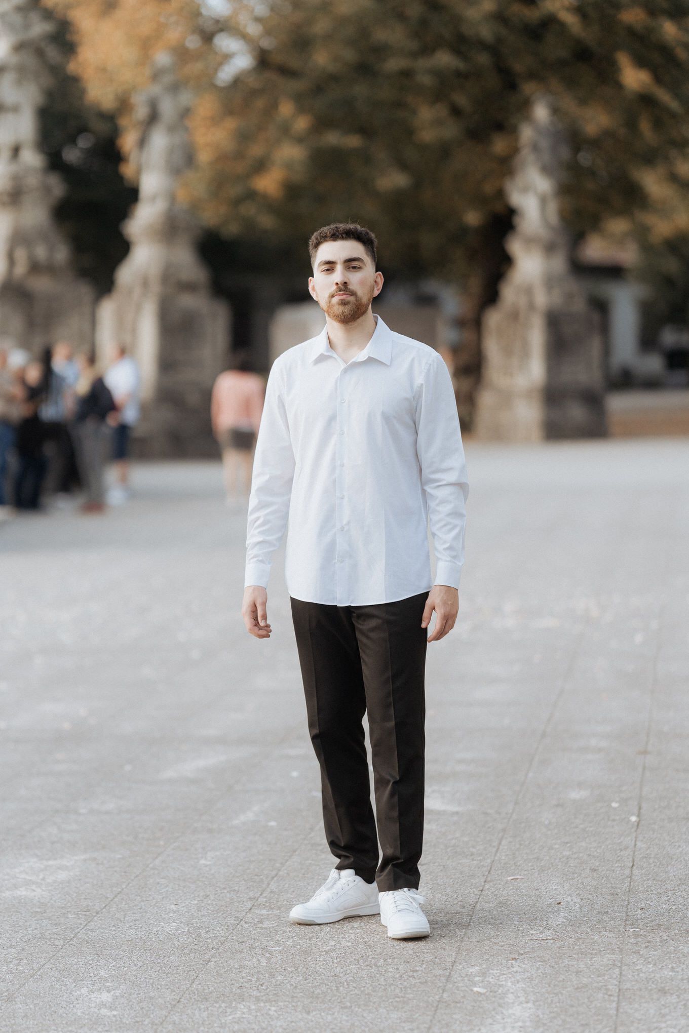 Retrato casual de um namorado vestindo uma camisa branca de botões e calças escuras, em pé em uma passarela de pedra no Bom Jesus de Braga.