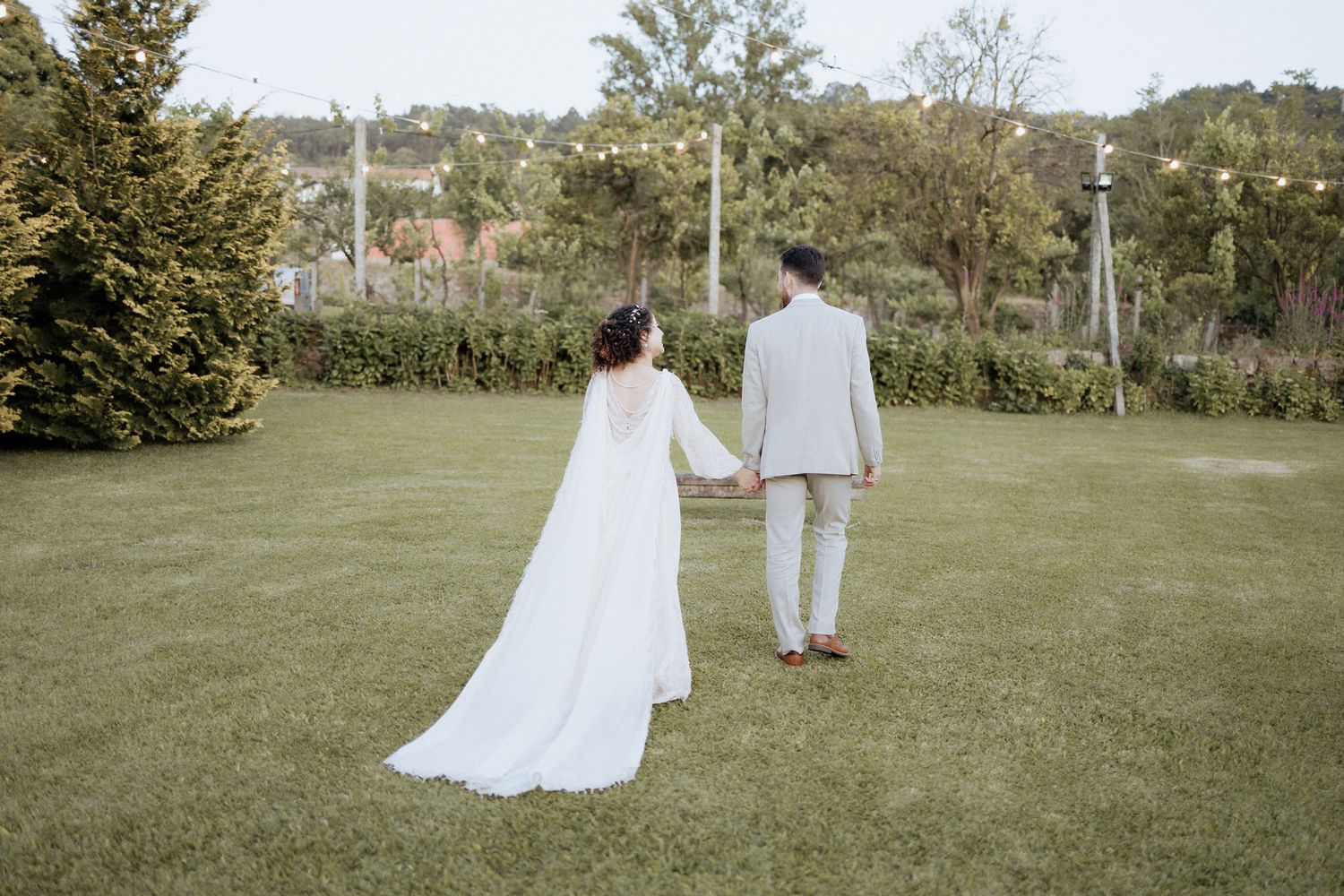 Couple in wedding attire walking on lush green lawn with trees in background during outdoor ceremony.