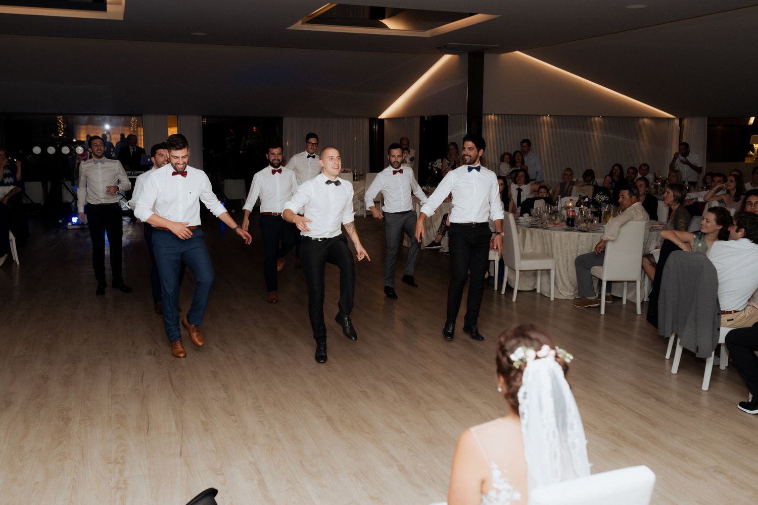 Groomsmen perform a coordinated dance line at a wedding reception on a wooden dance floor.