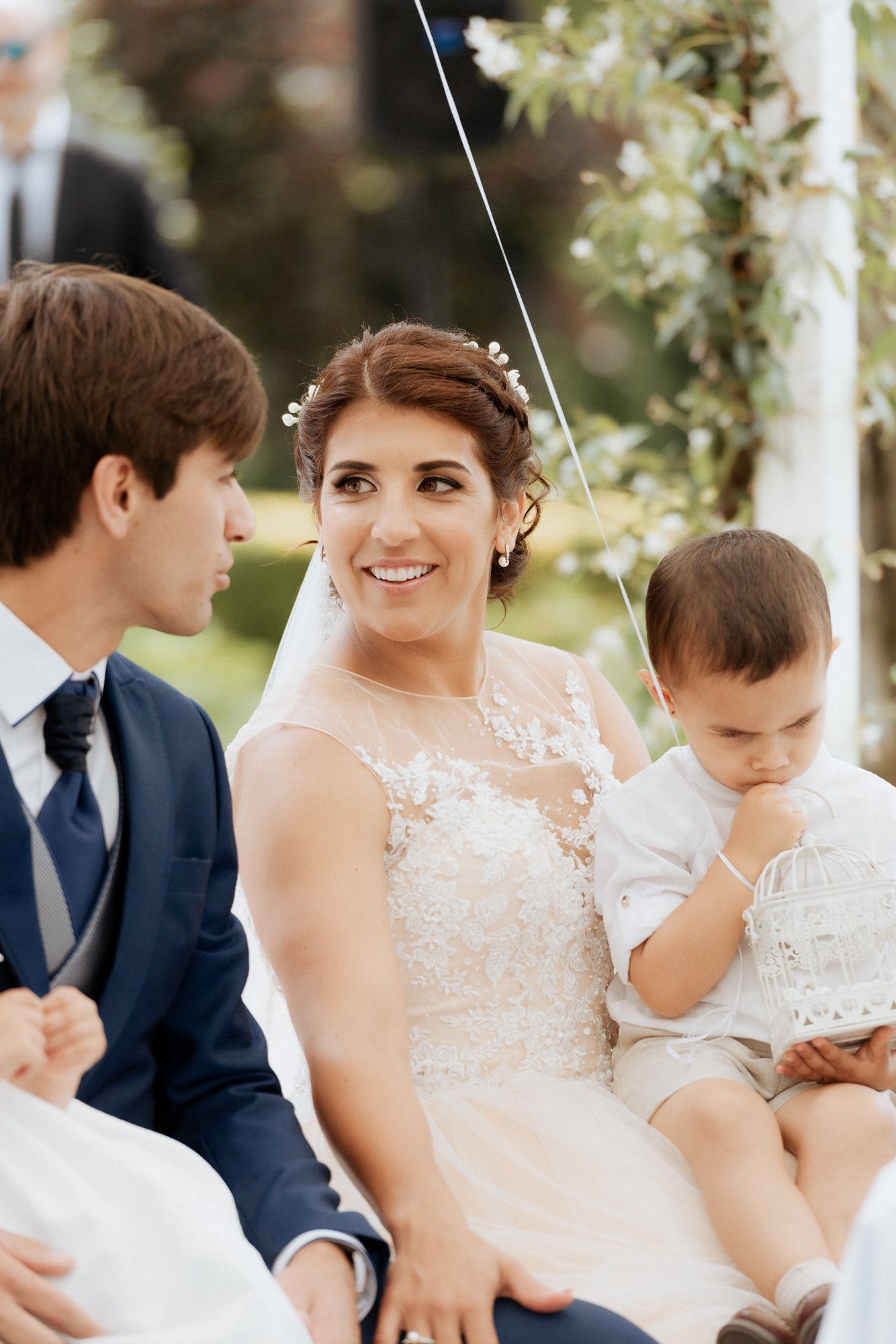 A couple on their wedding day sits outdoors, wearing elegant white attire and a navy suit.
