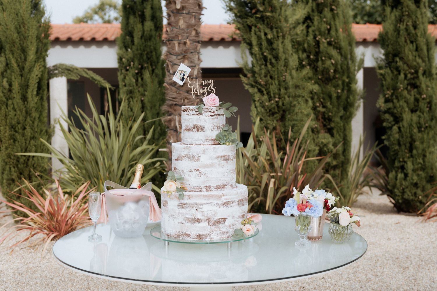 Elegant three-tiered semi-naked wedding cake displayed outdoors on glass table with modern desert garden backdrop.
