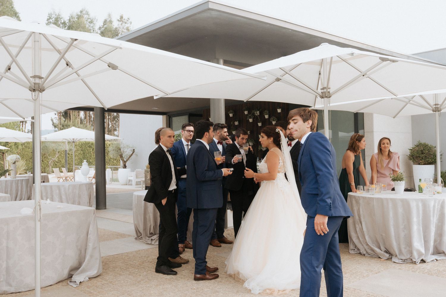 Wedding guests gather under white umbrellas at an outdoor reception with round tables and decorations at sunset.
