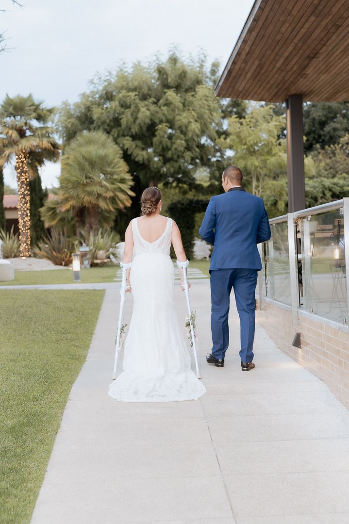 A couple in wedding attire walks together along a paved path beside a manicured lawn with palm trees in the background.