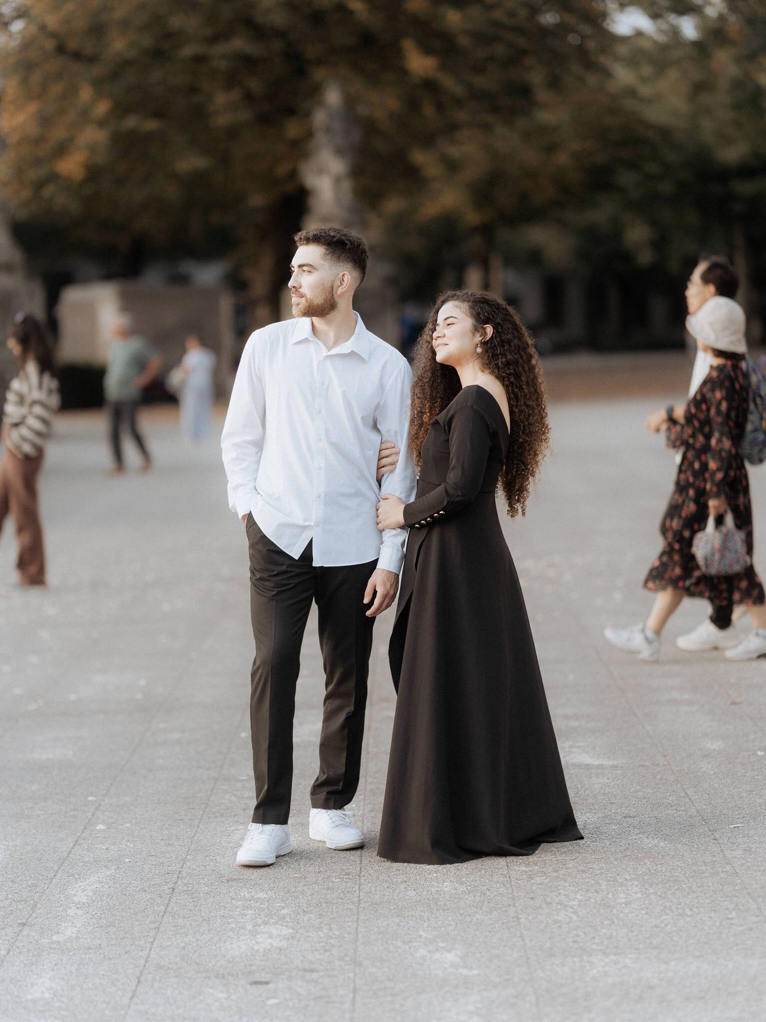 Um casal está junto ao ar livre, com uma pessoa de camisa branca e calça escura ao lado de outra com um vestido preto esvoaçante.