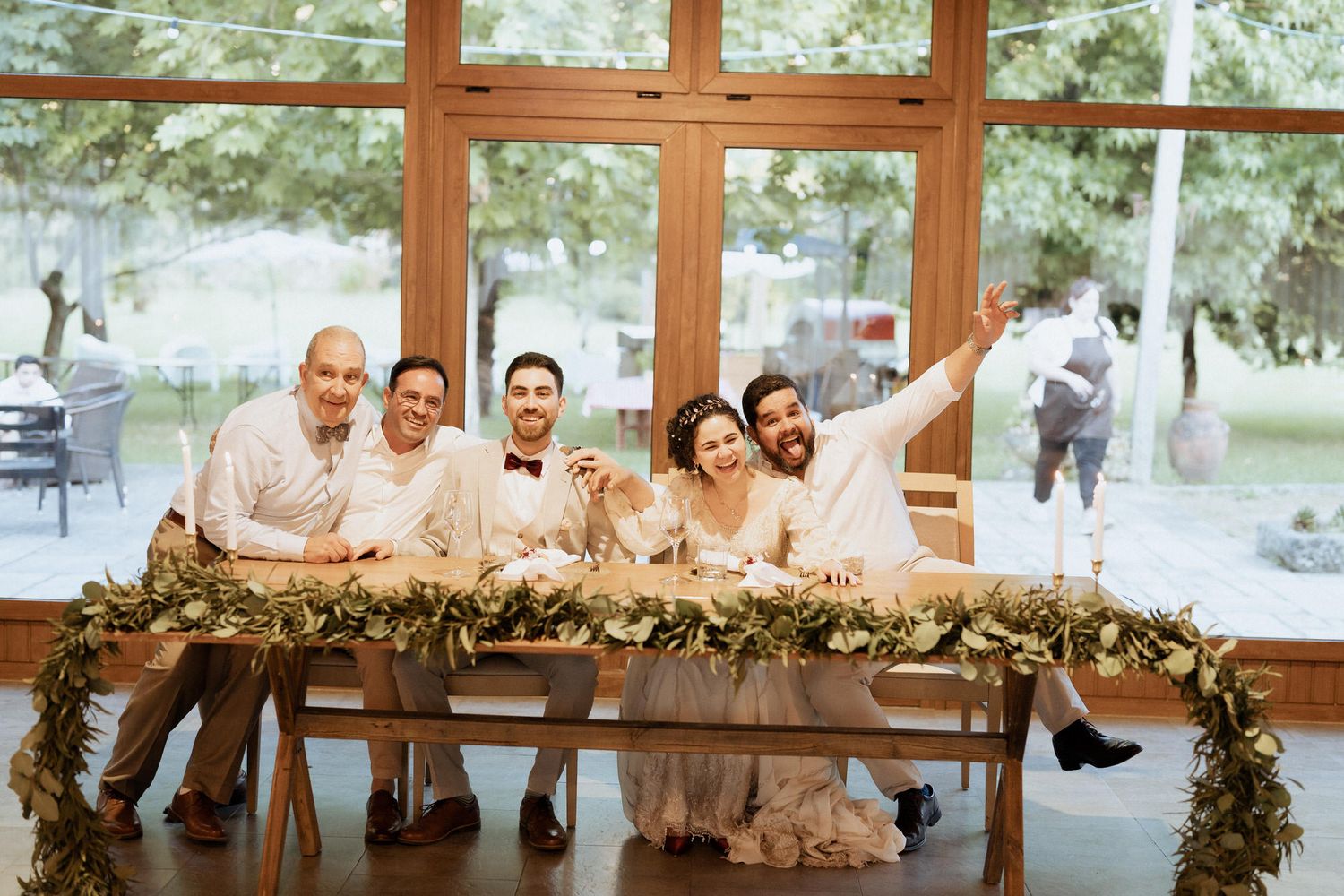 Wedding party in beige and cream attire pose together at a rustic wood table decorated with greenery garland.